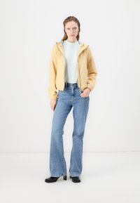 Giacca a vento gialla chiaro con cappuccio e polsini elasticizzati, abbinata a una maglietta verde menta e jeans a zampa d'elefante blu. Modella che indossa scarpe nere.