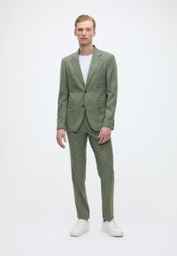 Grüner strukturierter Anzug mit zwei Knöpfen und fallenden Revers, kombiniert mit einem weißen Rundhalsausschnitt-T-Shirt und weißen Sneakers.