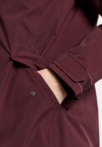 Burgunderfarbene wasserdichte Jacke mit seitlicher Tasche, Druckknopfverschluss und Klettverschluss-Akzenten an den Ärmelbündchen. Glatte, strukturierte Stoffoberfläche.