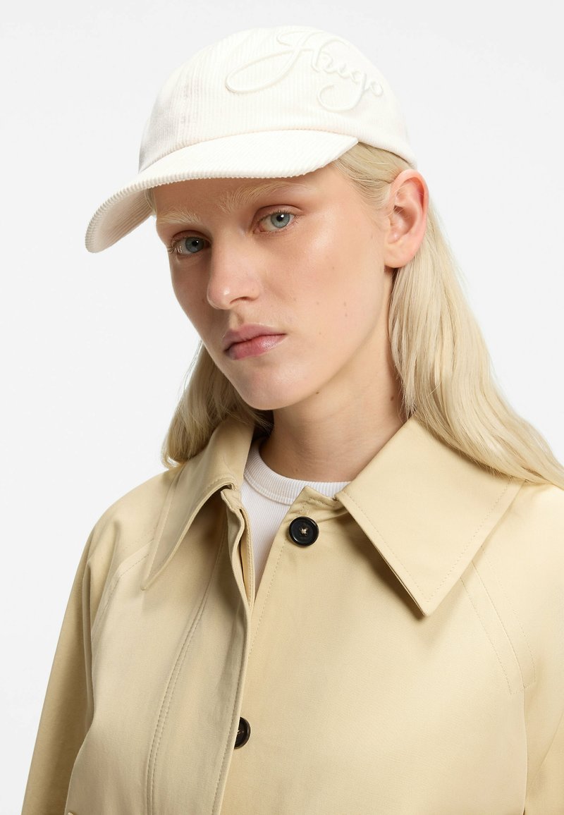 Femme blonde aux yeux bleus portant un trench-coat beige, une chemise blanche et une casquette de baseball crème avec un logo brodé.