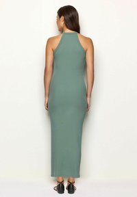 Femeie îmbrăcată cu o rochie fără mâneci, de culoare verde măsliniu, ajustată pe corp și până la gleznă, cu un spate tip racerback, purtând sandale negre cu brâu la gleznă, stând cu spatele.