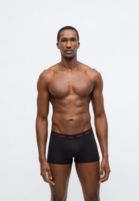 Μαύρα ανδρικά εσώρουχα boxer με κόκκινη μέση με λογότυπο, κατασκευασμένα από μαλακό ύφασμα. Ο σχεδιασμός περιλαμβάνει εφαρμογή που αγκαλιάζει το σώμα και κοντό μήκος ποδιού.