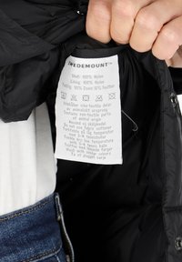 Inuti en svart nylonjacka med skötselinformation som beskriver material: 100% nylon yttertyg och foder, 90% dun, 10% fjäderfyllning.