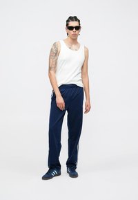 Camisola branca sem mangas, calças de treino azul-marinho com riscas laterais brancas e sapatilhas azuis. O modelo usa óculos de sol, exibindo tatuagens no braço esquerdo.