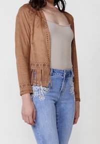 Femme portant une veste en daim beige avec des découpes florales et des franges, un débardeur beige et un jean bleu orné de motifs brodés blancs.