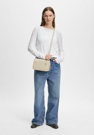 Frau trägt ein weißes Langarmshirt, weit geschnittene blaue Jeans, schwarze Schuhe und eine beigen Umhängetasche, steht vor einem schlichten weißen Hintergrund.