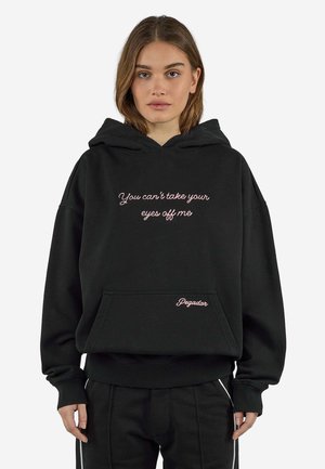 Ung kvinde med brunt hår bærer en sort hættetrøje med pink tekst "You can't take your eyes off me" og "Pegador" på forlommen.