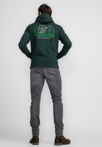 Sudadera verde oscura con un bolsillo frontal tipo canguro, con gráficos blancos de montañas y árboles en la espalda. Combinada con jeans gris entallados y botas marrones.