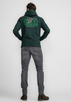 Sudadera verde oscura con un bolsillo frontal tipo canguro, con gráficos blancos de montañas y árboles en la espalda. Combinada con jeans gris entallados y botas marrones.