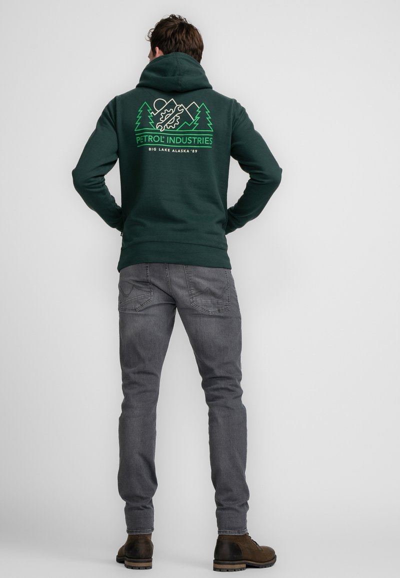 Sudadera verde oscura con un bolsillo frontal tipo canguro, con gráficos blancos de montañas y árboles en la espalda. Combinada con jeans gris entallados y botas marrones.