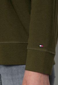 Camisa de manga larga color verde oliva con puños acanalados y un pequeño logotipo bordado en rojo, blanco y azul en el puño derecho. Textura suave.