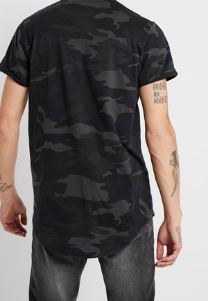 Osoba ubrana w czarną i ciemnoszarą koszulkę moro oraz ciemnoszare dżinsy, pokazująca tatuaże na obu przedramionach.