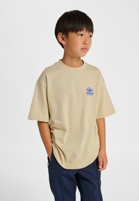 Beżowa koszulka z krótkim rękawem z niebieskim logo na lewej piersi. Odzież ma luźny krój i okrągły dekolt, zestawiona z ciemnymi spodniami.