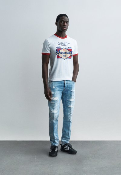 Άνδρας όρθιος με τα χέρια στις τσέπες, φορώντας λευκό T-shirt με κόκκινο περίγραμμα και γραφικά, ανοικτόχρωμο γαλάζιο τζιν με σκισίματα και μαύρα αθλητικά παπούτσια, μπροστά από απλό φόντο.