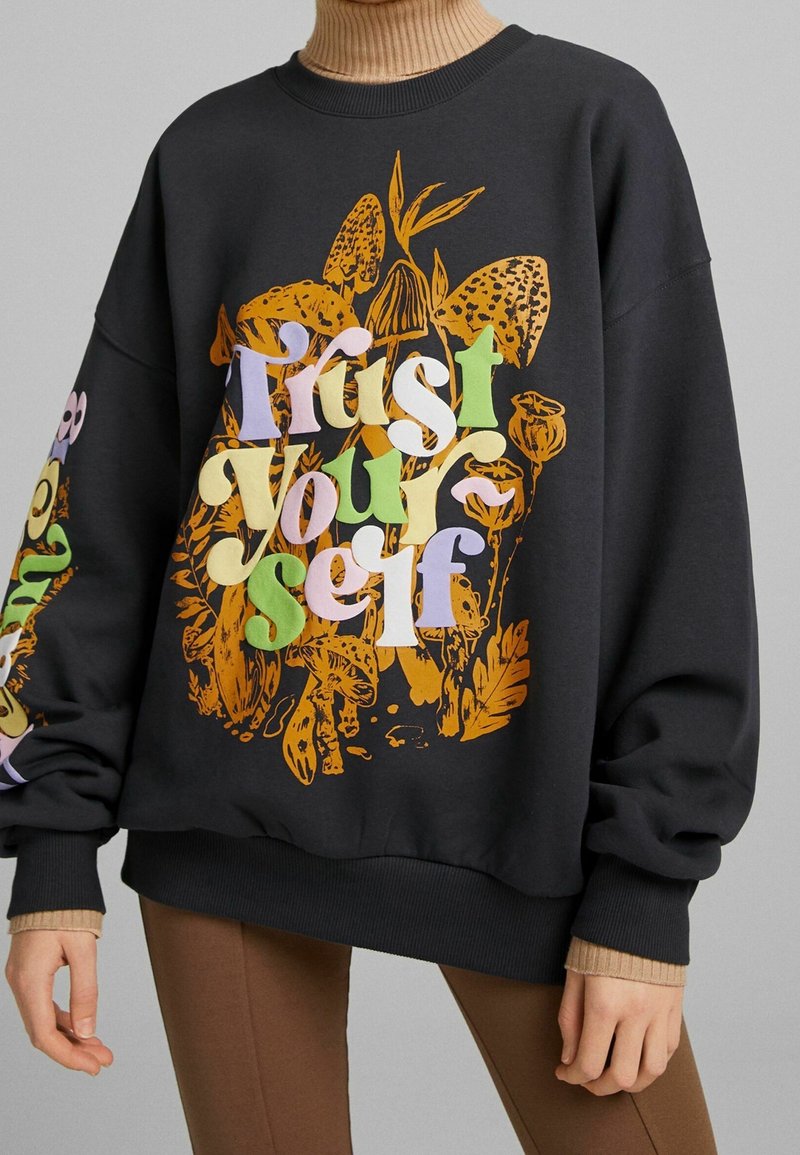 Personne portant un sweat-shirt noir avec « Fais-toi confiance » en lettres colorées sur un motif botanique orange, associé à un pantalon marron et un col roulé beige.