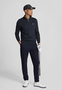Marineblauer Pullover mit Reißverschluss, grünes Hemd darunter, marineblaue Hose, weiße Golfschuhe, hält einen Golfschläger, trägt eine dunkle Kappe.