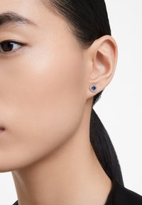 Brincos redondos de prata com uma pedra preciosa azul em forma de quadrado no centro, rodeada por um halo de pequenas pedras claras.