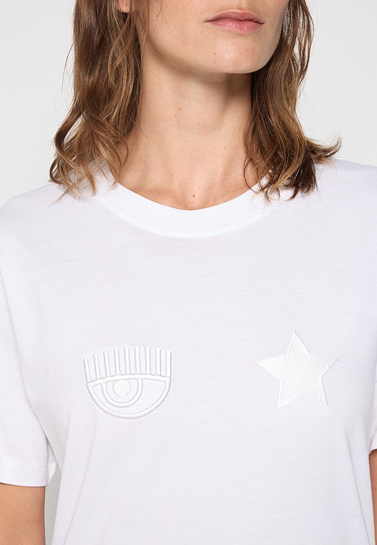 T-shirt en coton blanc avec des designs brodés d'un œil et d'une étoile sur le devant. Dispose d'un col rond classique et de manches courtes.