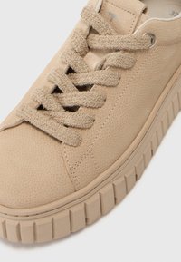 Beige sneakers med en teksturert skinnoverdel, flate tekstillisser og en tykk, rillede gummisåle. Har sølvfargede snorer og sømdetaljer.