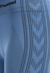 Nahaufnahme von blauem Sportstoff mit schwarzem, perforiertem Pfeilmuster und schwarzem "hummel"-Logo auf texturiertem Material.