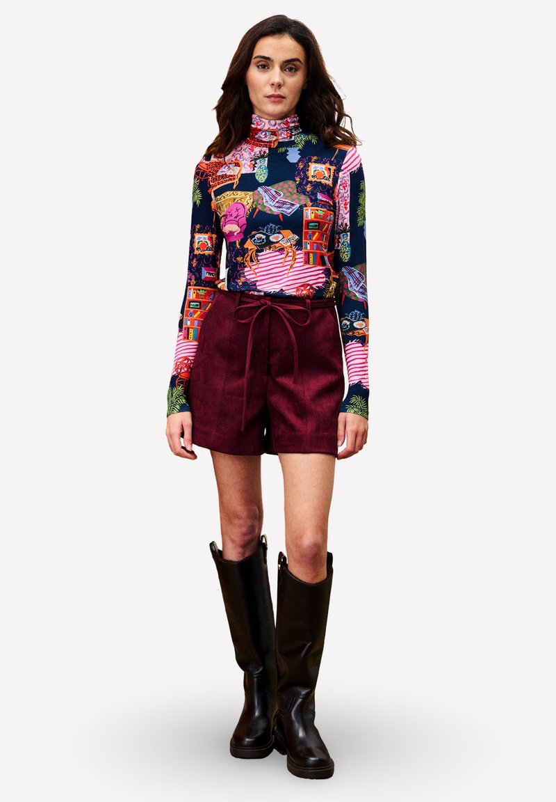 Frau in einem bunten, gemusterten Langarmrollkragenpullover, burgunderfarbenen, high-waist Shorts mit Bindegürtel und knielangen schwarzen Lederstiefeln, die steht.