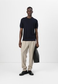 Mornarska kratka pletena jopica, bež karirani hlače, črne usnjene čevlje, drži črno usnjeno torbo. Preprost design, gladka tekstura tkanine.