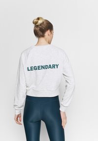 Šedá zkrácená mikina s nápisem "LEGENDARY" v tmavě zelené na zadní straně, doplněná lesklými teal legínami. Měkký materiál, volný střih.