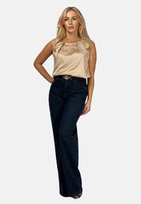 Beżowa satynowa bluzka bez rękawów z łańcuszkowym akcentem, zestawiona z ciemnoniebieskimi spodniami dzwonami o wysokim stanie, posiadającymi czarny pasek z złotą klamrą.