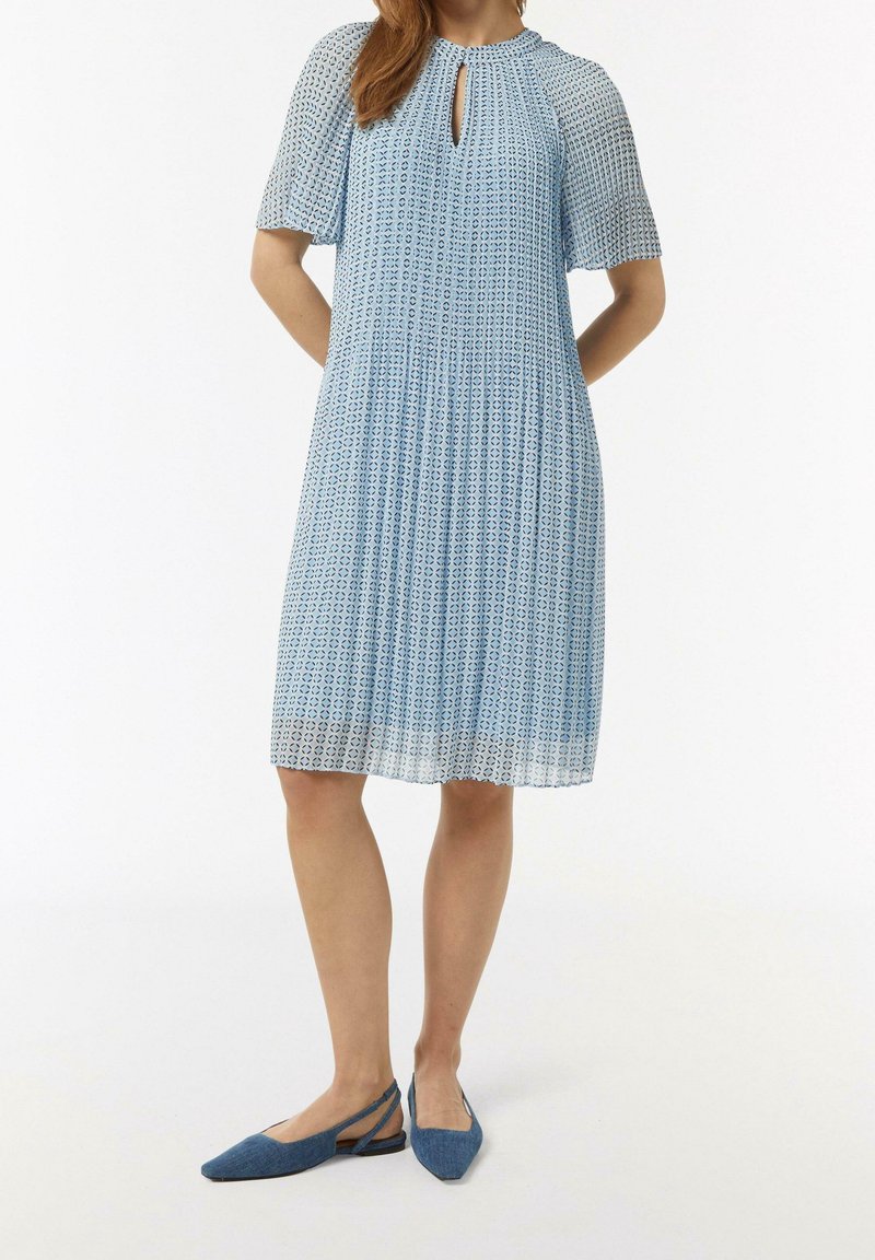 Femme portant une robe bleu clair mi-longue avec de petits motifs géométriques et des manches courtes, associée à des ballerines slingback bleues pointues.