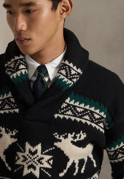 Μαύρο πουλόβερ με γιακά σαν σάλι, διακοσμημένο με λευκά και πράσινα πλεκτά μοτίβα ελαφιών και γεωμετρικά σχέδια. Φορεμένο πάνω από λευκό πουκάμισο και γραβάτα.
