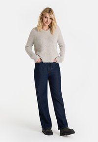 Hellgrauer gestrickter Pullover mit V-Ausschnitt und strukturiertem Muster, kombiniert mit dunkelblauen, weit geschnittenen Jeans und schwarzen Plate Stiefeln.