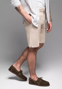Pantalones cortos beige con un acabado texturizado, cremallera frontal y aberturas laterales; combinados con zapatos de barco de ante marrón. Camisa de botones a rayas llevada abierta.