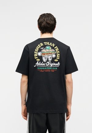 Persoon draagt een zwart Adidas Originals T-shirt met een cartoon van een sandwich, lopend, met tekst "Fresher Than Fresh" en stadsnamen op de achterkant.