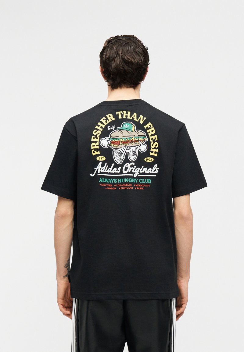 Άτομο που φοράει μαύρο T-shirt Adidas Originals με καρτούν σάντουιτς που περπατά, με το κείμενο «Fresher Than Fresh» και ονόματα πόλεων στην πλάτη.