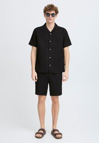 Cămașă neagră cu mâneci scurte și butoane, cu un model texturat, asortată cu pantaloni scurți. Ochelari de soare rotunzi și sandale maro completează ținuta.