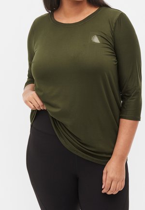 Oliven grøn, langærmet top med rund hals og et subtilt geometrisk design på venstre bryst. Blødt, strækbart stof. Sammen med sorte leggings.