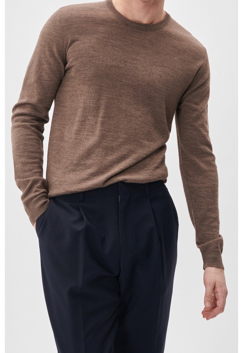 Homme portant un pull marron ajusté à manches longues et un pantalon plissé bleu marine foncé, debout avec une main dans la poche sur fond uni.