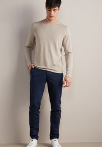 Beżowa koszula z długim rękawem w połączeniu z granatowymi spodniami slim-fit i białymi sneakersami. Tkanina wydaje się miękka, gładka i lekka. Brak widocznych wzorów.