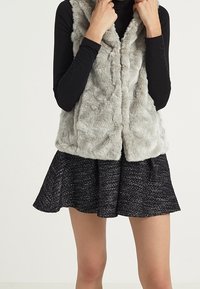 Grå fuskpälsväst med hög krage, bärs över en svart långärmad topp, ihop med en svart kjol med en texturerad mönstring.