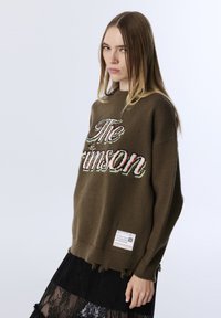 Brauner Strickpullover mit buntem Stickmotiv "The Crimson", lässiger Schnitt und fransigen Kanten. Etikett an der unteren Seite mit Pflegehinweisen.