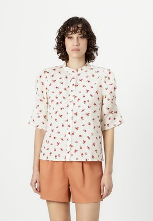 Bloemige overhemdd met korte pofmouwen, witte stof, met een klein rood bloemenpatroon en een ronde halslijn. Gecombineerd met oranje shorts.