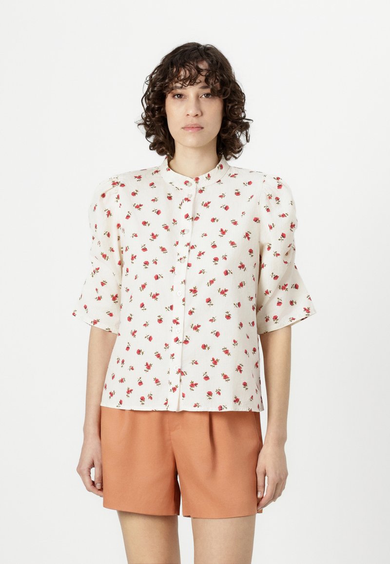 Floral button-up shirt with short puff sleeves, white fabric, small red flower print, and a round neckline. Paired with orange shorts.