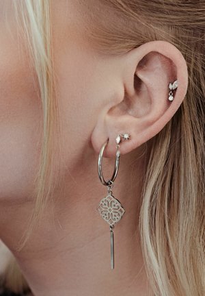 Close-up of a left ear with four silver earrings, including a large hoop with a patterned charm and three small stud and drop designs.