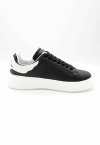 Zapatilla baja negra con suela blanca gruesa, cordones negros y detalle blanco en forma de ala en el talón, vista lateral sobre fondo blanco.