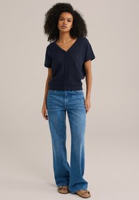 Tums zila adīta krekla forma ar pogām, V-veida kaklu un īsām piedurknēm, kopā ar gaiši ziliem platām kājām džinsiem un brūnām sandalēm. Gluda tekstūra, ikdieniška piegriezums.