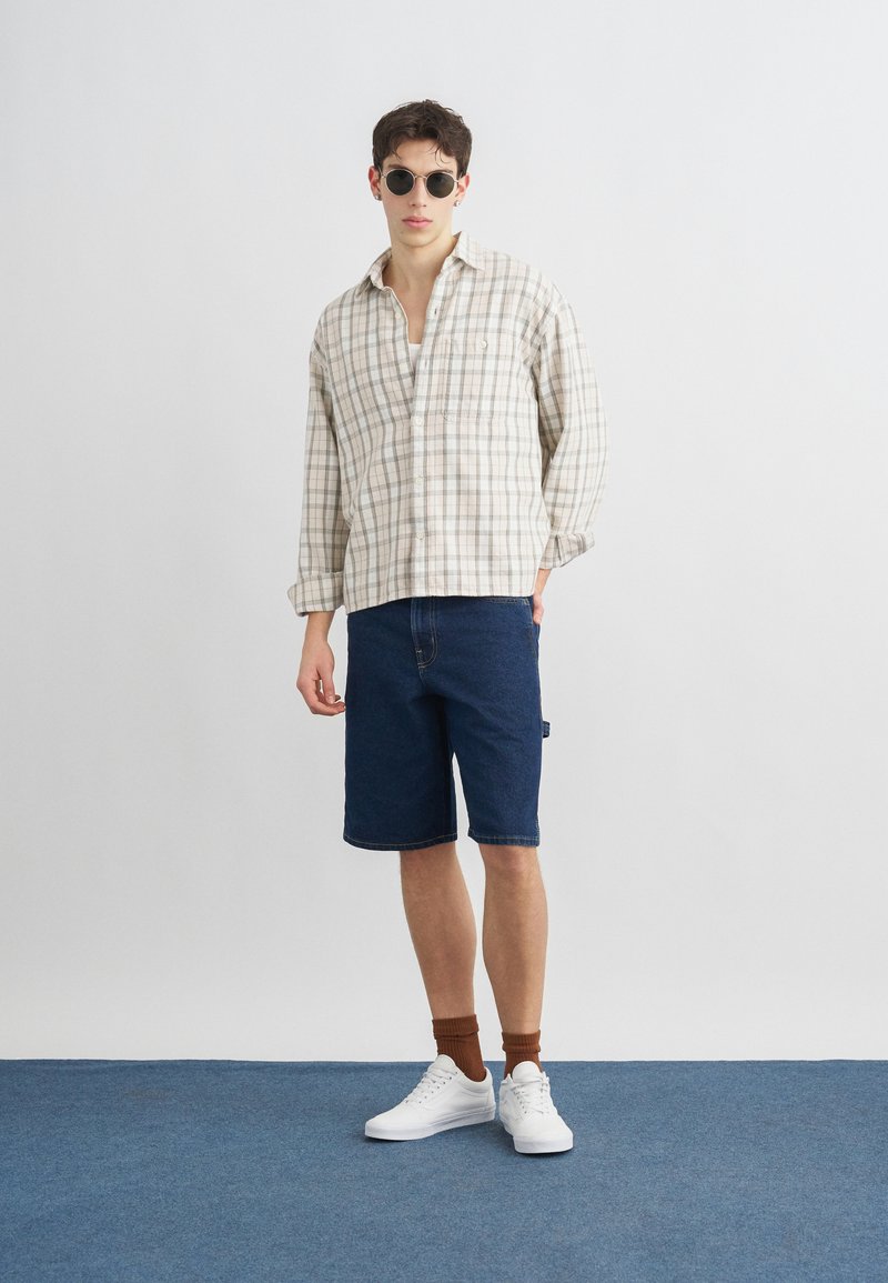 Giovane uomo con occhiali da sole rotondi, camicia beige a quadri con bottoni, pantaloncini di jeans blu scuro, calzini marroni e sneakers bianche, in piedi su un tappeto blu.