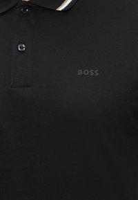Polo nera con due bottoni, colletto a coste con righe bianche e beige e logo "BOSS" ricamato sul petto.