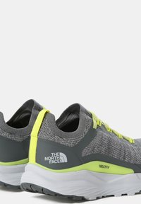 Zapatillas deportivas grises con parte superior de malla texturizada, acentos en amarillo neón y media suela blanca, que presentan un logotipo destacado y una pestaña de tiro.