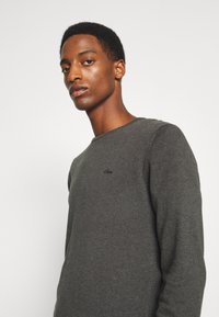 Dunkelgraues Langarmshirt aus einem weichen, strukturierten Stoff mit Rundhalsausschnitt und einem kleinen gestickten Logo auf der Brust.