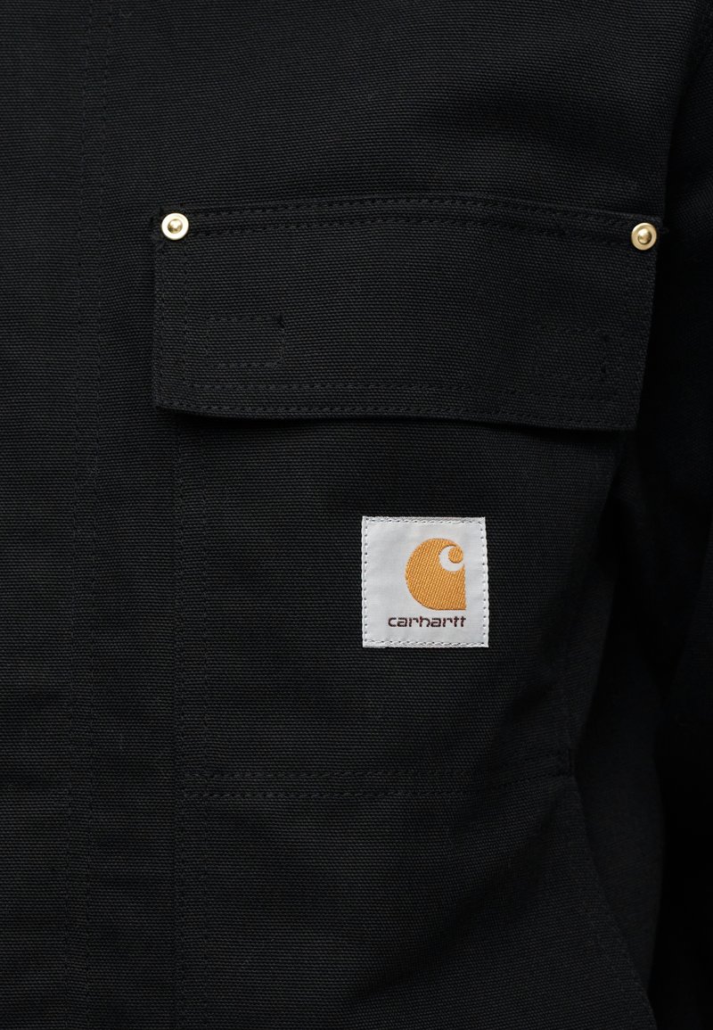 Zwart stoffen overhemd met twee borstzakken. Opvallende details zijn onder andere goudkleurige hardware en een vierkante witte label met een oranje logo.